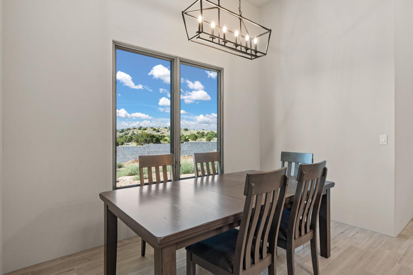 Dining table with wooden chairs by large windows showing a bright outdoor view.