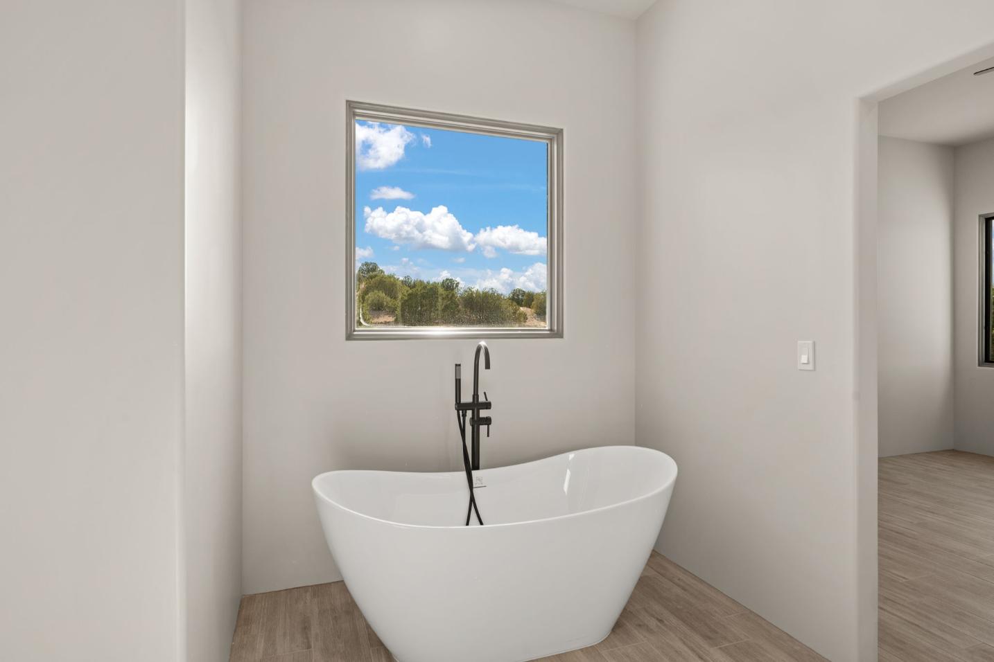 Modern freestanding bathtub under a window with a scenic view of clouds and trees.