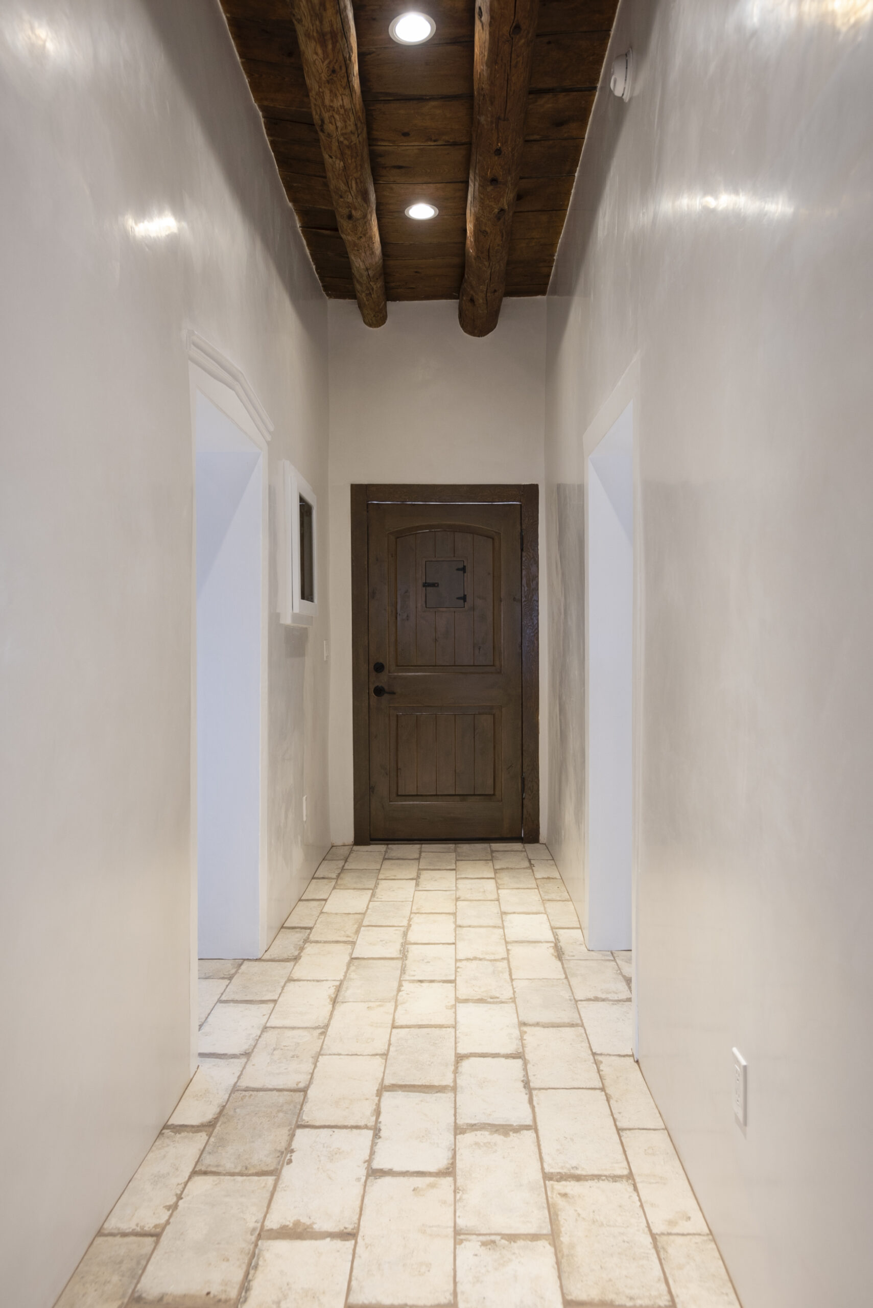 A narrow hallway with white walls, tiled floor, and a wooden door at the end.